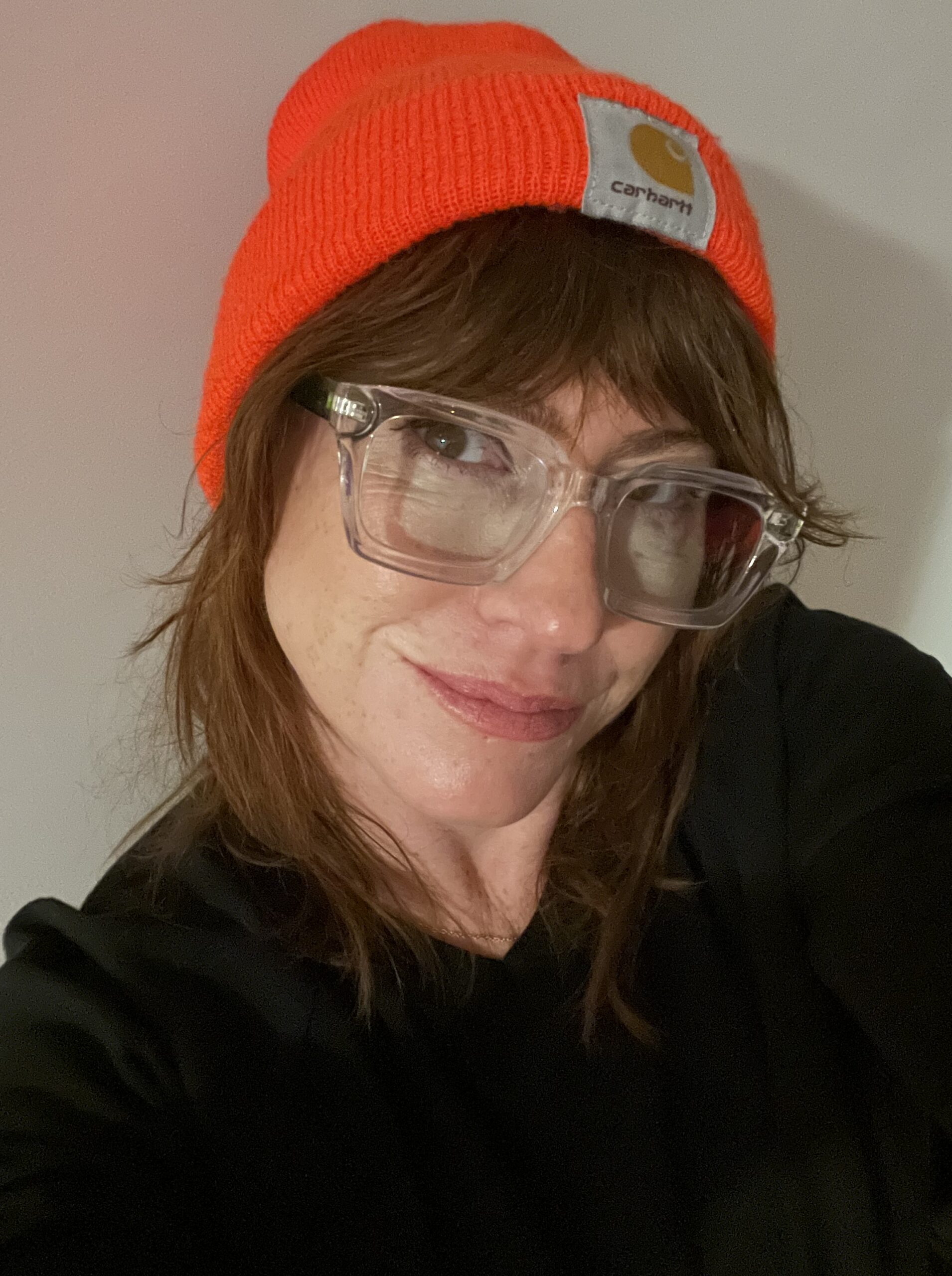 Genderqueer Menopause author, Lasara Firefox Allen, in a bright orange beanie, black shirt, and clear glasses.